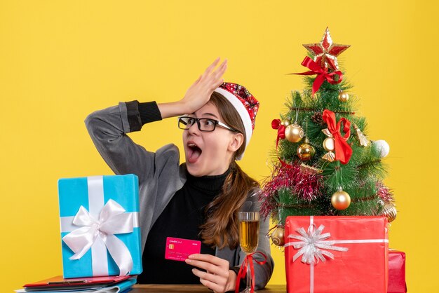 Front view wide-eyed girl with xmas hat sitting at the table putting hand