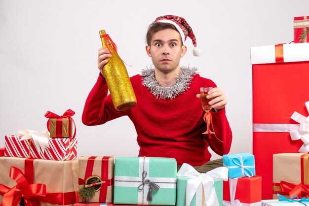 Front view wide-eyed young man with santa hat holding champagne sitting around xmas gifts