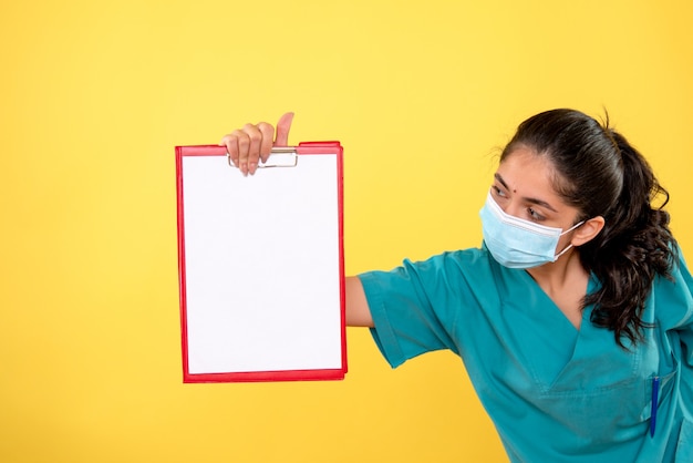 Free Photo front view of woman doctor in uniform looking at clipboard standing on yellow wall