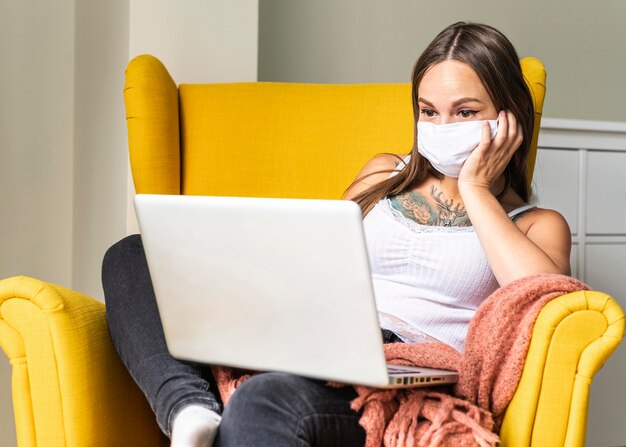 Front view of woman with medical mask working on laptop from armchair during the pandemic