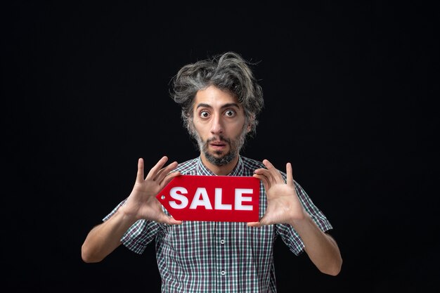 Front view of wondered man holding sale sign on dark wall