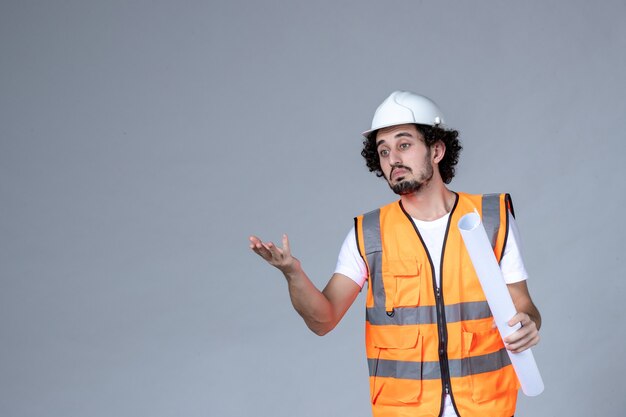 Front view of wondering male constructor in warning vest wearing safety helmet and holding blank pointing forward on gray wave wall