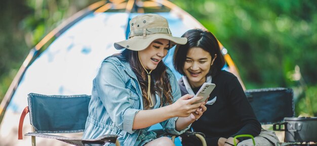 Front view Young Asian pretty woman and her girlfriend sitting at front of tent use mobile phone take photo during camping in forest with happiness together