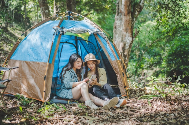 Free photo front view young asian pretty woman and her girlfriend sitting at front of tent use mobile phone take photo during camping in forest with happiness together