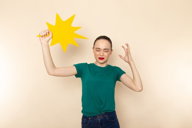 Front view young attractive female in dark green shirt and blue jeans holding yellow sign on beige