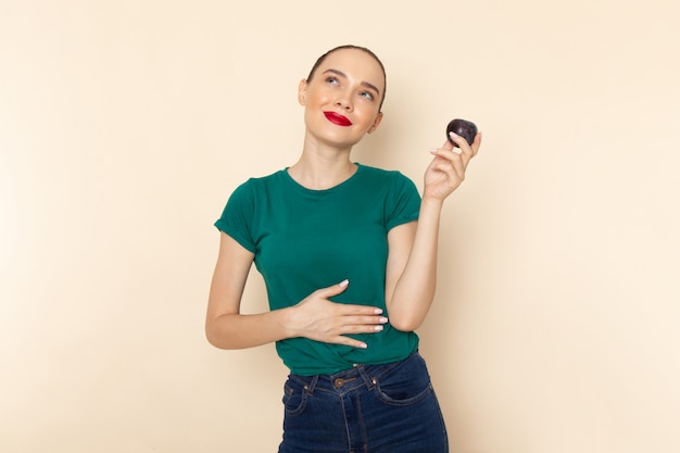 Front view young attractive female in dark green shirt holding plum and smiling on beige