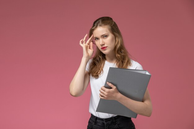 Front view young attractive female in white t-shirt holding grey file with thinking expression on the pink desk model female color female young