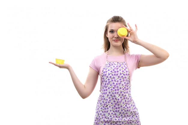 A front view young attractive housewife in pink shirt colorful cape holding yellow thing smiling on the white background cuisine kitchen female