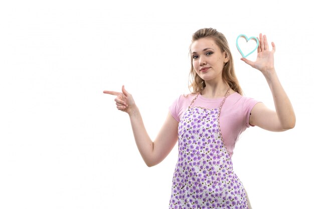 A front view young attractive housewife in pink shirt colorful cape posing smiling holding little blue heart shape on the white background cuisine kitchen female