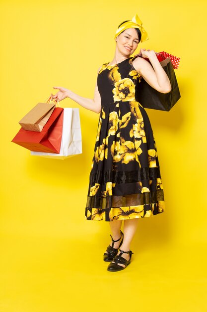 A front view young attractive lady in yellow-black flower designed dress with yellow bandage on head smiling holding shopping packages on the yellow