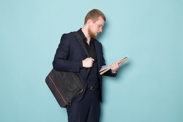 Free photo a front view young attractive male with beard in black dark classic modern suit holding files backpack on the blue space