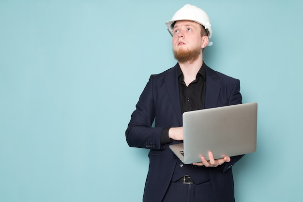 Free photo a front view young attractive male with beard in black dark classic modern suit white construction helmet using grey laptop on the blue space