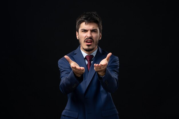 Front view of young bearded man in suit asking something with suspicious facial expression on isolated dark wall