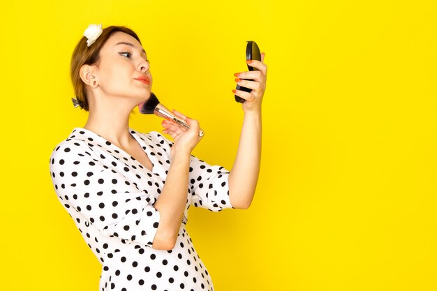 A front view young beautiful female in black and white polka dot dress doing make-up on yellow