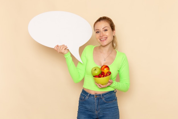 Free Photo front view young beautiful female in green shirt holding plate full of fruits and white sign on cream wall fruit model woman food vitamine color