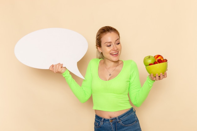 Front view young beautiful female in green shirt holding plate full of fruits with a big white sign on cream wall fruit model woman