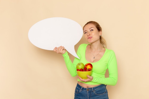 Free Photo front view young beautiful female in green shirt holding plate full of fruits with white sign on the cream wall fruit model woman food vitamine color