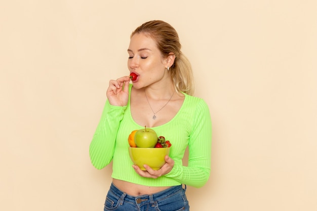 Front view young beautiful female in green shirt holding plate with fruits and eating strawberry on light cream wall fruit model woman pose