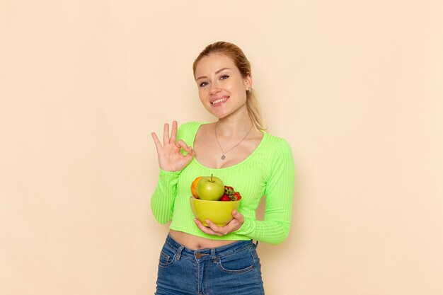 Front view young beautiful female in green shirt holding plate with fruits smiling on light cream floor fruit model woman pose lady