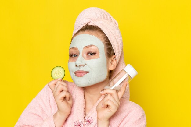 A front view young beautiful female in pink bathrobe holding spray and cucumber slice