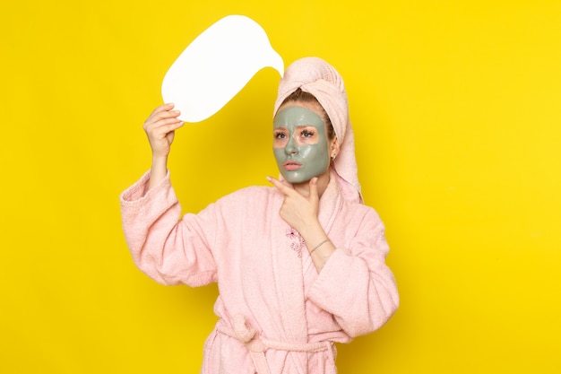 A front view young beautiful female in pink bathrobe with face-mask holding white sign thinking