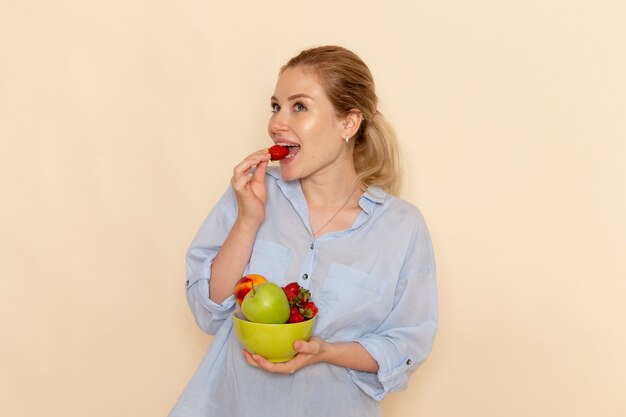 Front view young beautiful female in shirt holding plate with fruits eating strawberry on the cream wall fruit ripe model woman pose