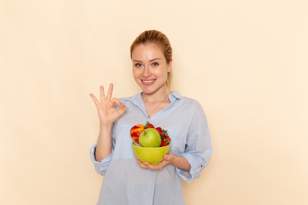 Free photo front view young beautiful female in shirt holding plate with fruits showing alright sign on the cream wall fruit ripe model woman pose