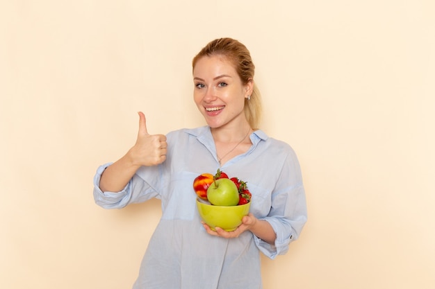 Free photo front view young beautiful female in shirt holding plate with fruits with smile on the cream wall fruit ripe model woman pose