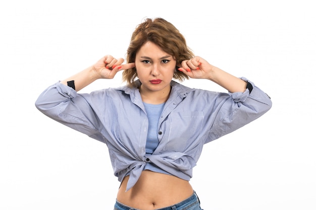 A front view young beautiful girl in blue shirt and blue jeans covering her ears on the white