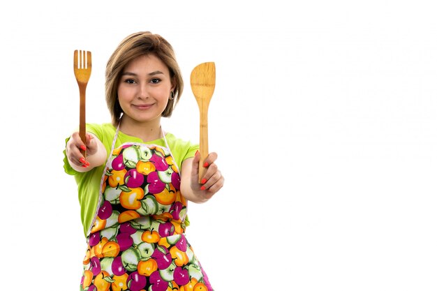 A front view young beautiful housewife in green shirt colorful cape holding wooden kitchen appliance smiling on the white background house cleaning kitchen