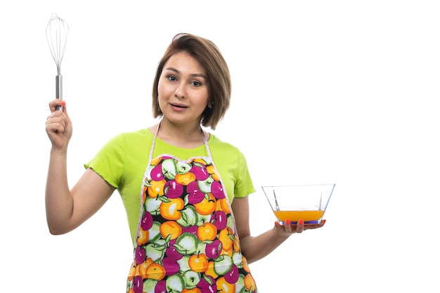 A front view young beautiful housewife in green shirt colorful cape mixing liquid eggs preparing meal on the white background house female kitchen