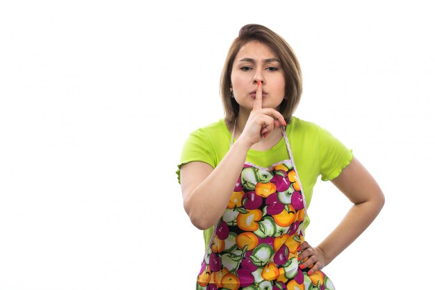 A front view young beautiful housewife in green shirt colorful cape posing showing silence sign on the white background house female kitchen