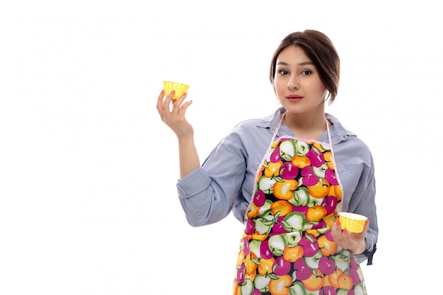 A front view young beautiful lady in light blue shirt and colorful cape holding yellow cake pans