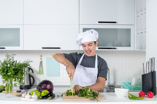 Front view young chef in uniform cutting greens on cutting board in kitchen