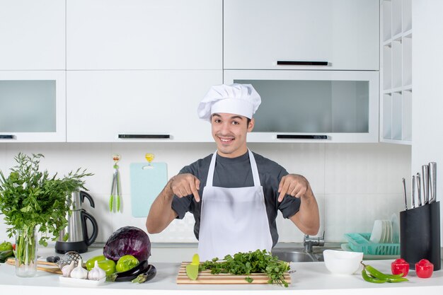 Front view young chef in uniform pointing at greens on cutting board