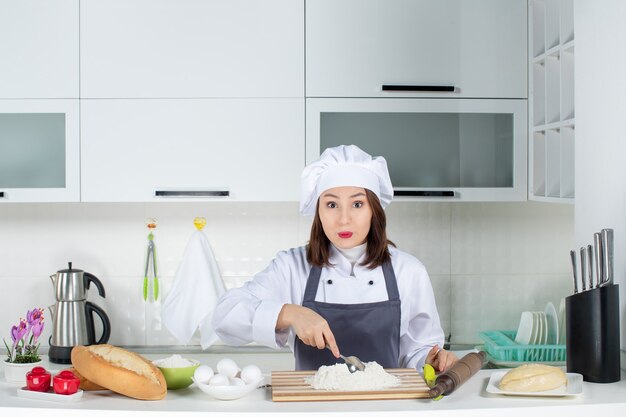 Front view of young confident female chef in uniform preparing food in the white kitchen