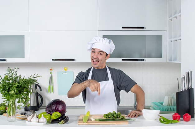 Front view young cook in uniform pointing at green on cutting board