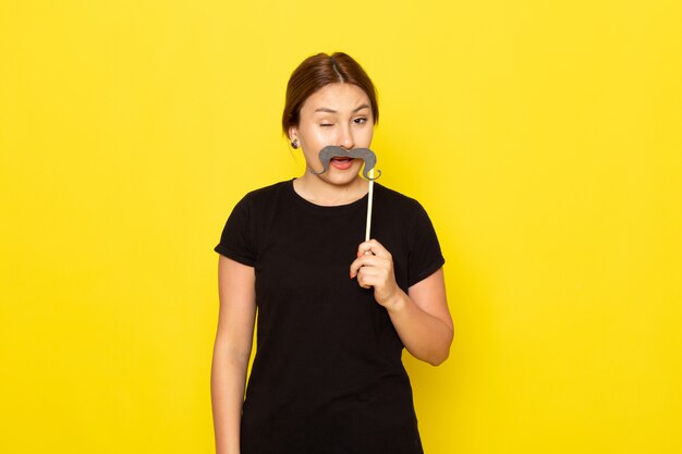 A front view young female in black dress posing with fake mustache with funny expression on yellow