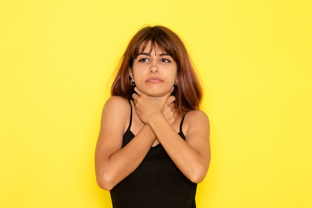 Front view of young female in black shirt and grey jeans chocking herself on yellow wall