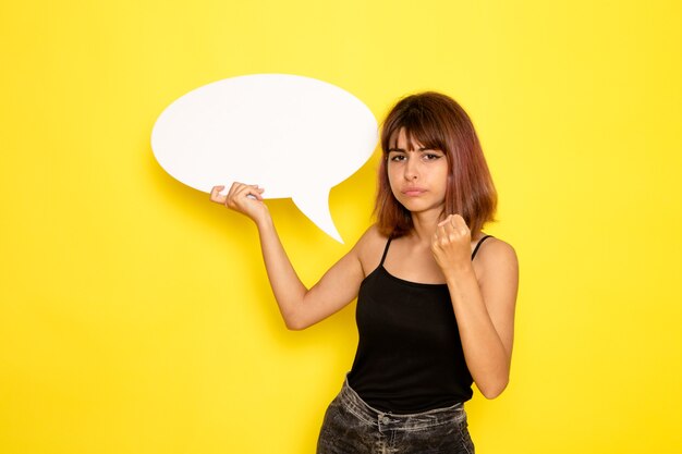 Front view of young female in black shirt and grey jeans holding big white sign on yellow wall