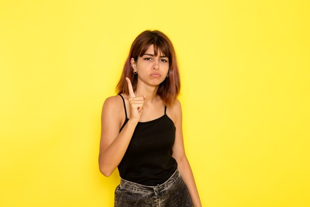 Front view of young female in black shirt and grey jeans threatening with her finger on yellow wall