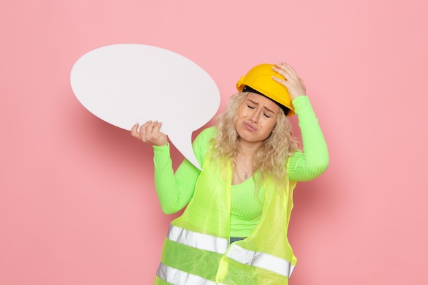 Free photo front view young female builder in green construction suit helmet holding white sign depressed on the pink space