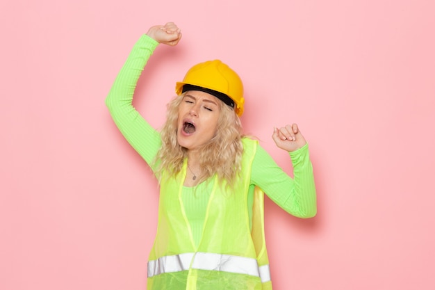 Front view young female builder in green construction suit helmet yawning on the pink space architecture construction work lady job