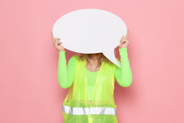 Free Photo front view young female builder in green construction suit yellow helmet holding white big sign on the pink space architecture construction job