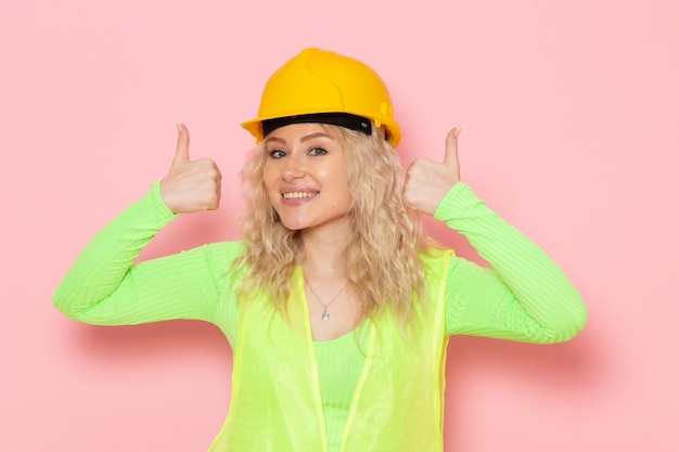 Free photo front view young female builder in green construction suit yellow helmet just posing with smile on the pink space architecture construction job