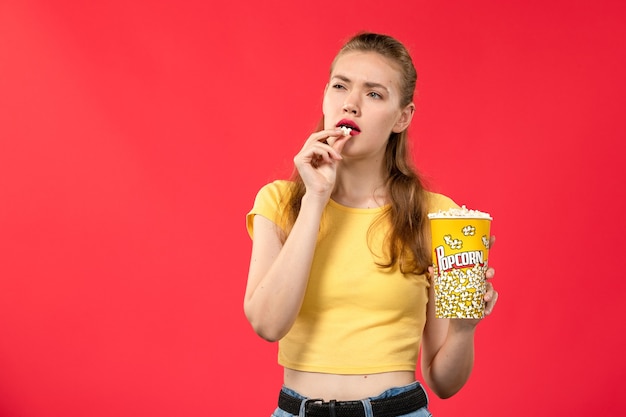 Front view young female at cinema holding and eating popcorn package on the light-red wall movie theater cinema female fun film