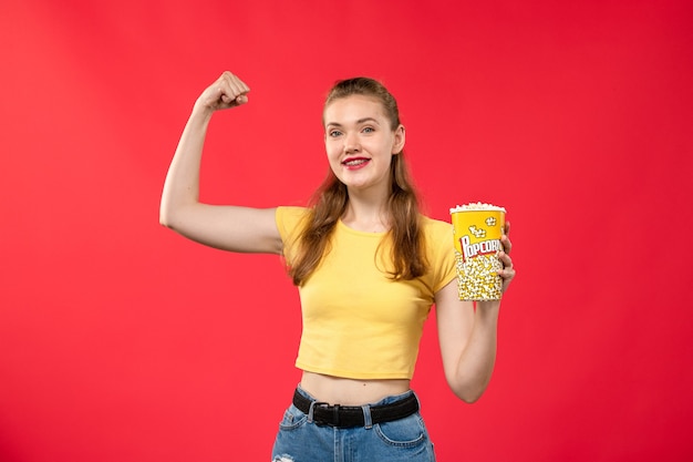 Free photo front view young female at cinema holding popcorn package and flexing on red wall movies theater cinema snack female fun film