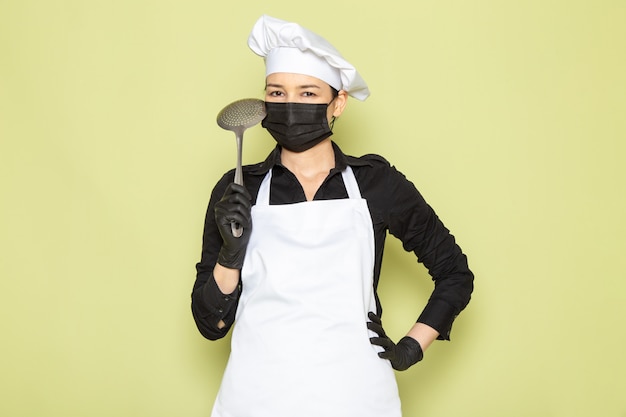 Free photo a front view young female cook in black shirt white cook cape white cap in black gloves black mask posing holding big silver spoon