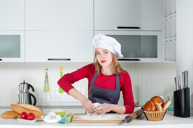 Front view young female cook in cook hat and apron kneading dough in the kitchen