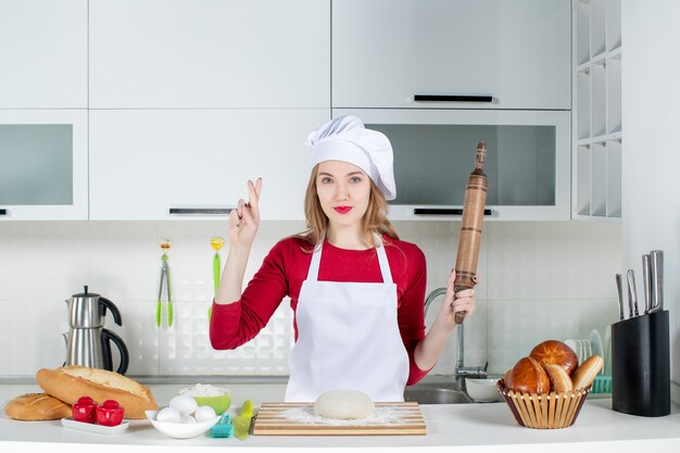 Front view young female cook holding rolling pin making good luck sign in the kitchen